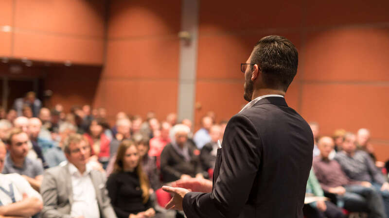 homme devant une salle pleine de gens en plein discours