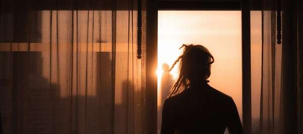 Une femme de dos devant une fenêtre au lever du soleil, illustrant le temps qu'il faut pour aller mieux après une rupture