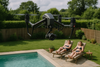 Drone noir survolant un jardin clôturé avec une piscine, tandis que deux personnes bronzent au loin sur des transats et regardent le drone d’un air mécontent.
