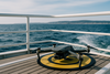Drone de loisirs en catégorie Open posé sur une zone de décollage circulaire jaune sur le pont d’un bateau en pleine mer, face à l’océan Atlantique légèrement agité, avec une côte au loin sous un ciel bleu voilé
