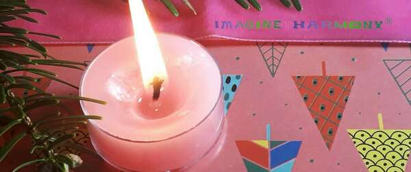 Une bougie rose parfumée à la rose est allumée. A côté d'elle, se trouvent une branche de sapin et un ruban rose. Le tout est posé sur des dessins de sapins décolrés sur fond rose.