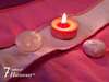 Cette photo montre une composition zen et apaisante avec une bougie rouge allumée posée sur un tissu aux tons roses et mauves, accompagnée de deux pierres polies (une rose claire et une mauve). 