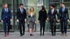 6 business persons walk towards the lens lens. The 2 business women are at the center of the group.