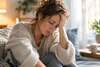 Femme assise chez elle, la tête penchée et la main sur le front, évoquant la fatigue et l’épuisement.