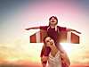 Une femme qui tient son enfant sur les épaules, et l'enfant est vêtu d'un déguisement d'aviateur avec des ailes et il essaie de s'envoler